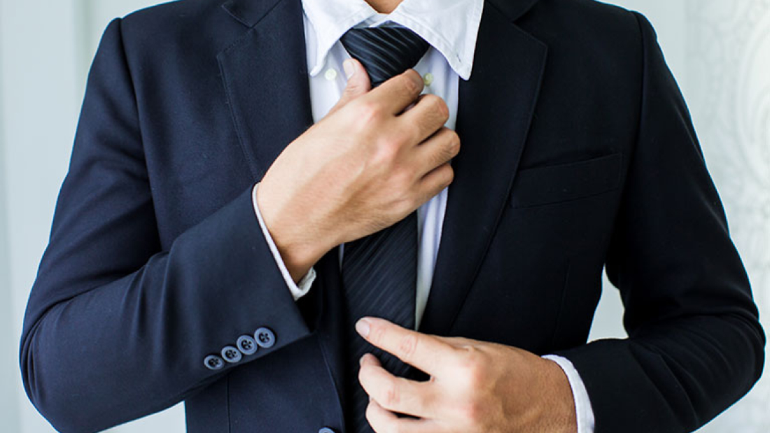 Zu sehen ist der Torso eines Mannes in dunkelblauem Anzug, der seine Krawatte richtet. 