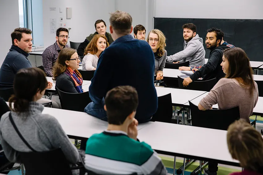 Dozent sitzt umringt von Student:innen in einem Seminarraum.