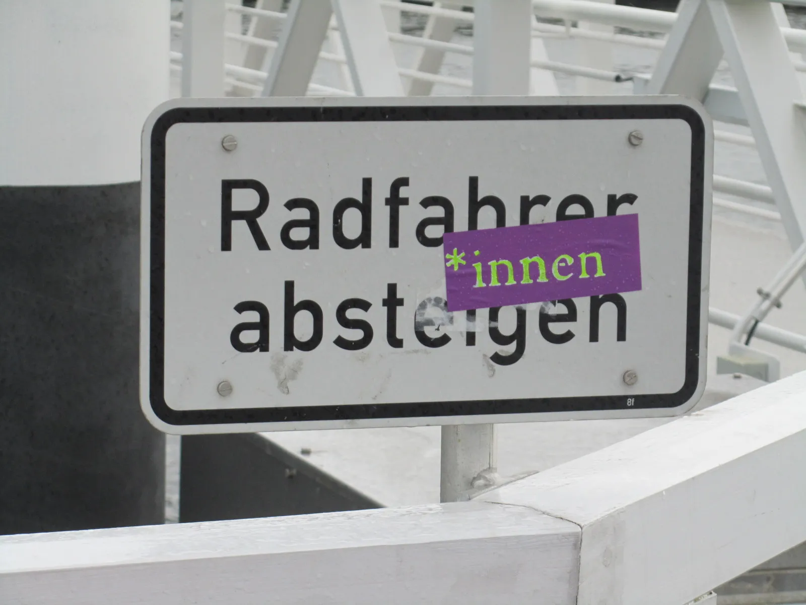 Ein Schild ist zu sehen, das "Radfahrer absteigen" besagt, auf dem ein Aufkleber mit der Aufschrift "*innen" angebracht wurde.