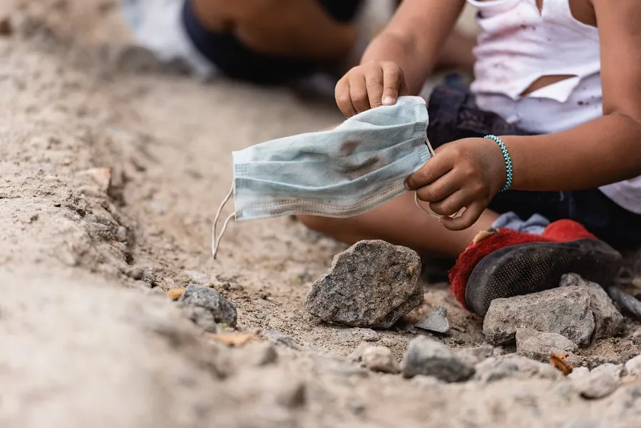 Man sieht Hände eines Kindes, das auf der Erde zwischen Steinen sitzt und eine schmutzige medizinische Maske hält.