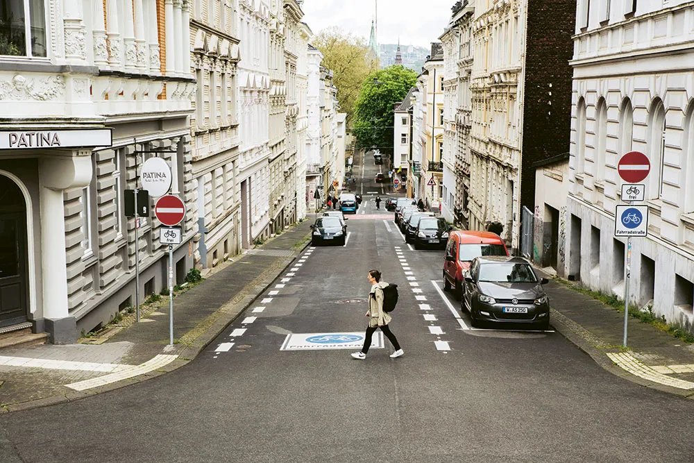 Frau geht von rechts nach links über eine Straße, links und rechts stehen Häuser