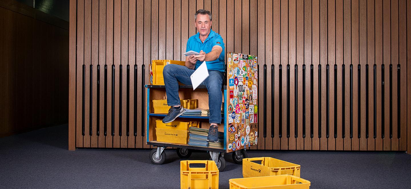 Mann auf Wägelchen vor getäfelter Wand wirft Dokumente in Post-Boxen