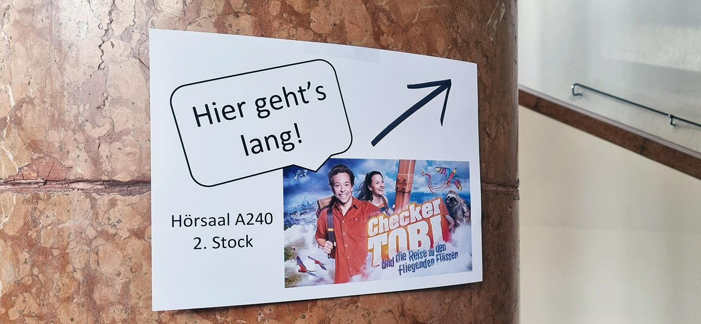 Schild mit "Hier geht's lang" und einem Kinoplakat in einem Treppenhaus
