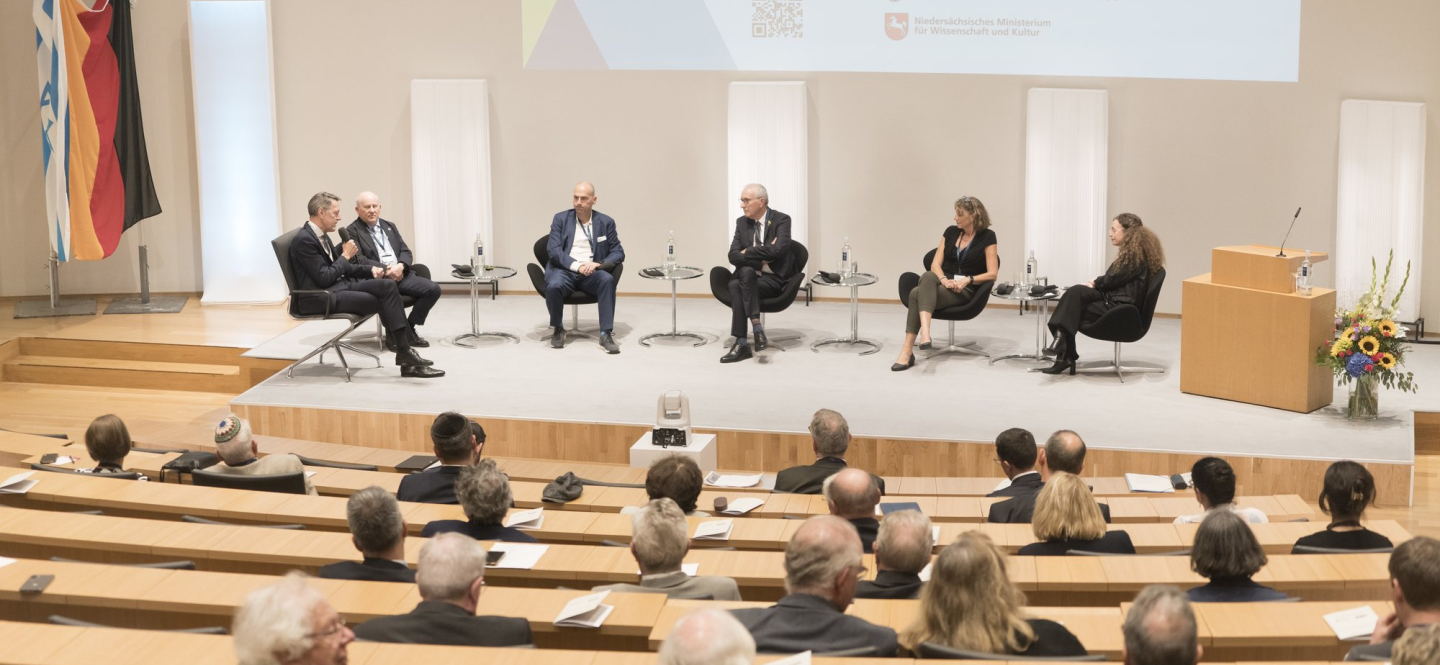 Podiumsdiskussion mit sechs Teilnehmer:innen, die nebeneinander auf einer Bühne sitzen
