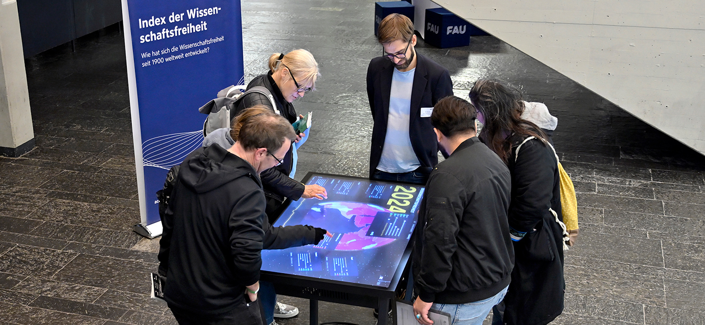 mehrere Personen stehen um einen flachen Bildschirm, auf dem eine in verschiedenen Farben eingefärbte Weltkarte zu sehen ist, dahinter steht ein Banner mit der Aufschrift "Index der Wissenschaftsfreiheit"