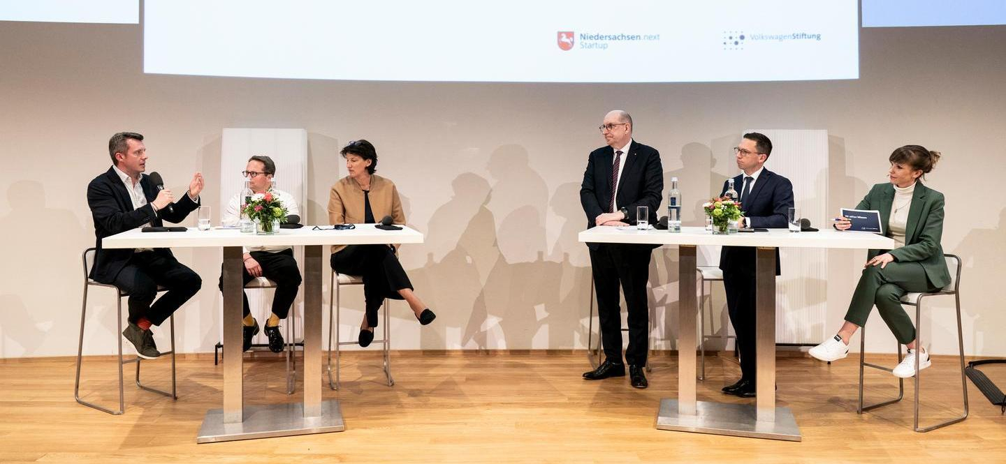 Sechs Menschen stehen an zwei Stehtischen auf einer Bühne und sprechen miteinander