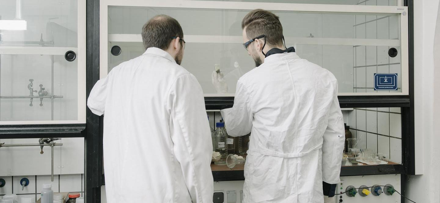 Zwei Männer im Kittel stehen vor einem Abzug im Chemielabor und betrachten eine Probe