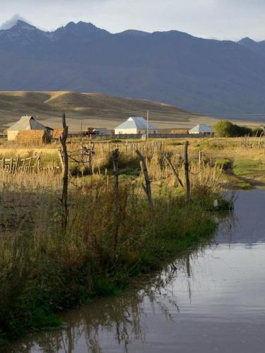 Bach vor einzelnen Häusern in weiter Landschaft, im Hintergrund hohe Berge