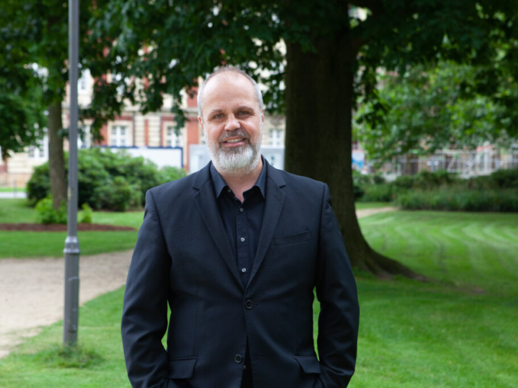 Das Bild zeigt einen lächelnden Mann in einem Park stehen.