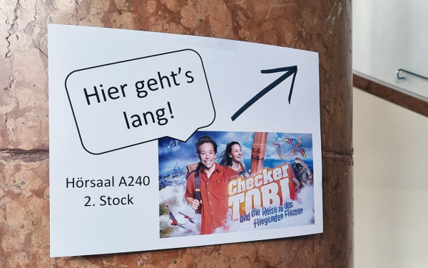 Schild mit "Hier geht's lang" und einem Kinoplakat in einem Treppenhaus