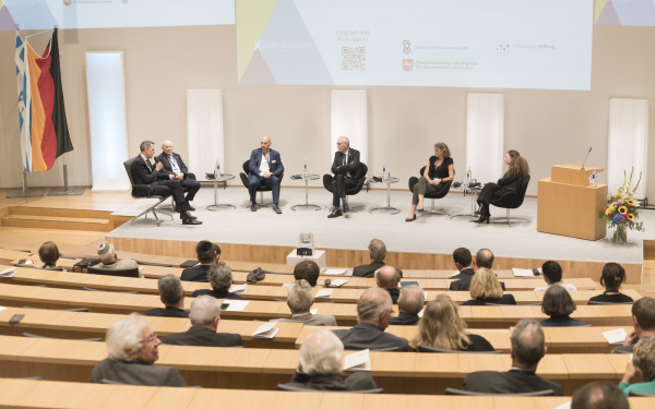 Podiumsdiskussion mit sechs Teilnehmer:innen, die nebeneinander auf einer Bühne sitzen