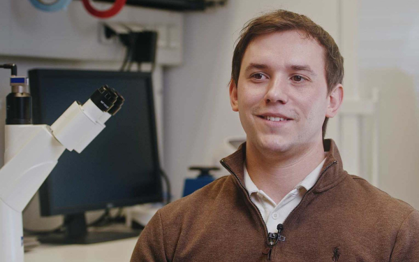 Ein Mann im braunen Pullover sitzt neben einem Mikroskop in einem Labor