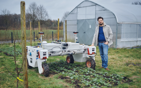 Ein Mann steht recht neben einem weißen Roboter auf vier Rädern auf einem Feld 