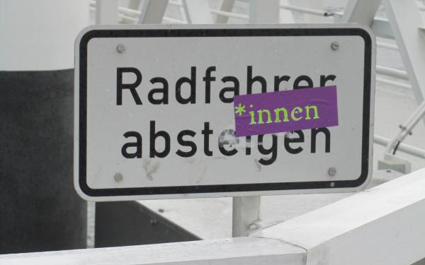 Ein Schild ist zu sehen, das "Radfahrer absteigen" besagt, auf dem ein Aufkleber mit der Aufschrift "*innen" angebracht wurde.