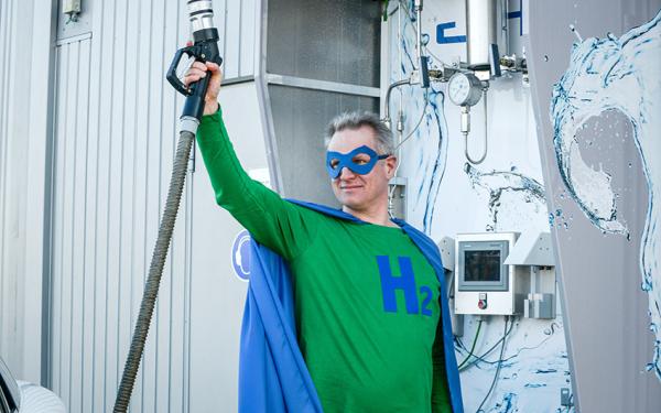 Richard Hanke-Rauschenbach trägt ein Heldenkostüm mit einem "H2"-Eblem und hält einen Tankrüssel für Wasserstoff in die Luft.