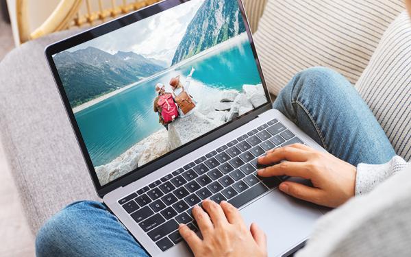 Person hat einen Laptop auf dem Schoß, auf dem man ein Bild mit zwei Personen am Bergsee sieht