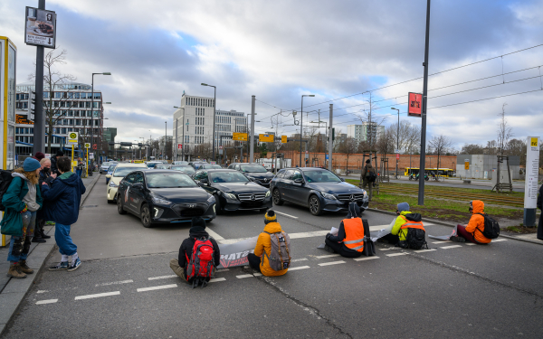 Das Bild zeigt Personen, die auf einer Straße sitzen und die Durchfahrt der Autos blockieren.