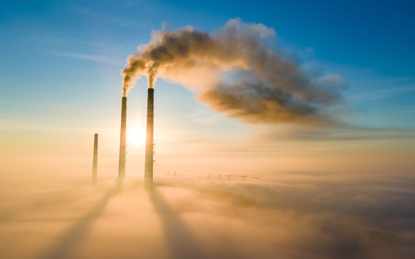 Das Bild zeigt drei industrielle Schornsteine, die Abgase vor einem Sonnenuntergang ausblasen.ist die EU-Flagge zu sehen,