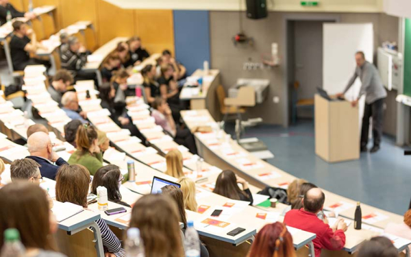 Das Bild zeigt einen Hörsaal mit Studierenden in den Reihen und einem Dozenten am Pult