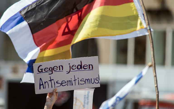 Das Bild zeigt eine Hand, die ein Pappschild hochhält. Darauf steht geschrieben "Gegen jeden Antisemitismus". Im Hintergrund sieht man die deutsche und die israelische Flagge.