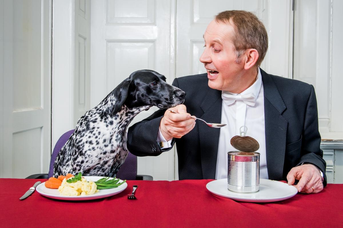 Mann im Anzug und ein Hund speisen zusammen an einem Tisch, der Hund sitzt hinter einem Teller Gemüse, der Mann hinter eine geöffneten (Hundefutter?) Dose
