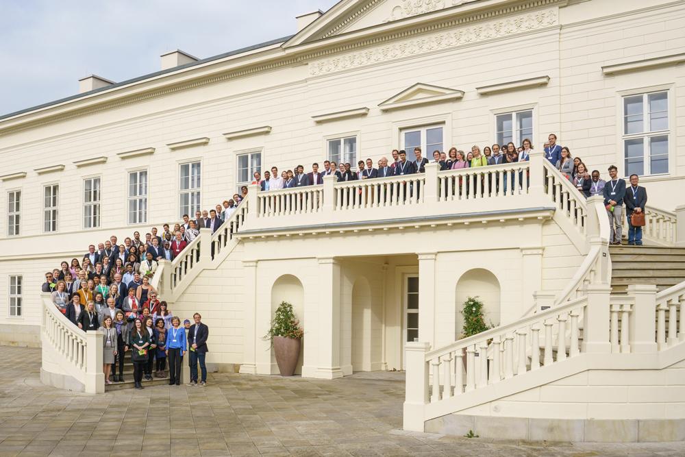 Menschen stehen auf der Außentreppe des in beige gehaltenen Schlosss Herrenhausen und posieren für ein Gruppenfoto.