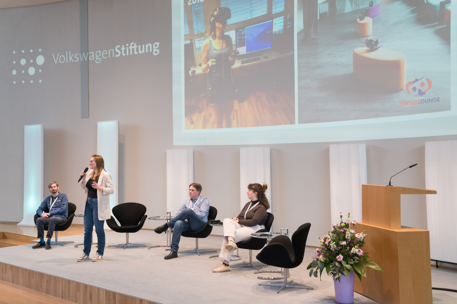 Eine Frau präsentiert einen Vortrag mit Mikro in der Hand, drei weitere Menschen sitzen mit ihr auf dem Podium