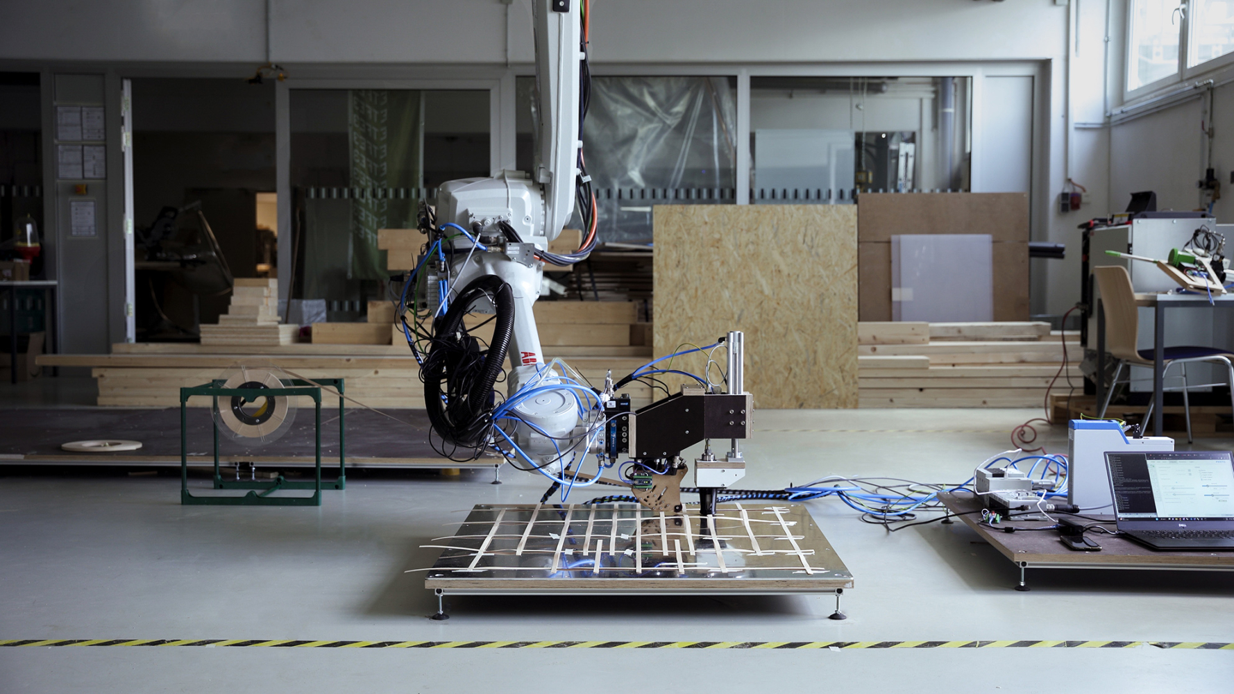 Ein Roboterarm ist in einer Laborumgebung zu sehen. Er schweißt Holzstreifen auf eine große Platte.