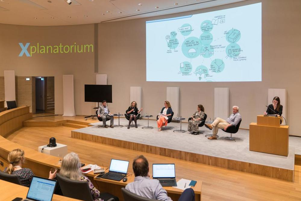 Podiumsdiskussion mit fünf Teilnehmenden vor Publikum, rechts zeichnet eine Frau eine Sketch note zum Gesagten, die im Hintergrund an die Wand projiziert wird. 