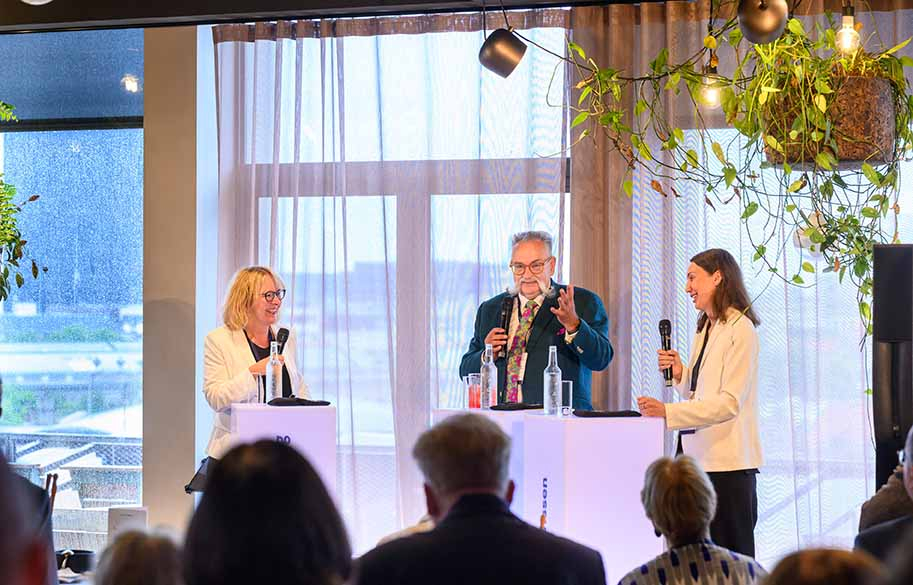 Drei Personen stehen an Pulten und halten Mikros in den Händen, während sie miteinander sprechen.