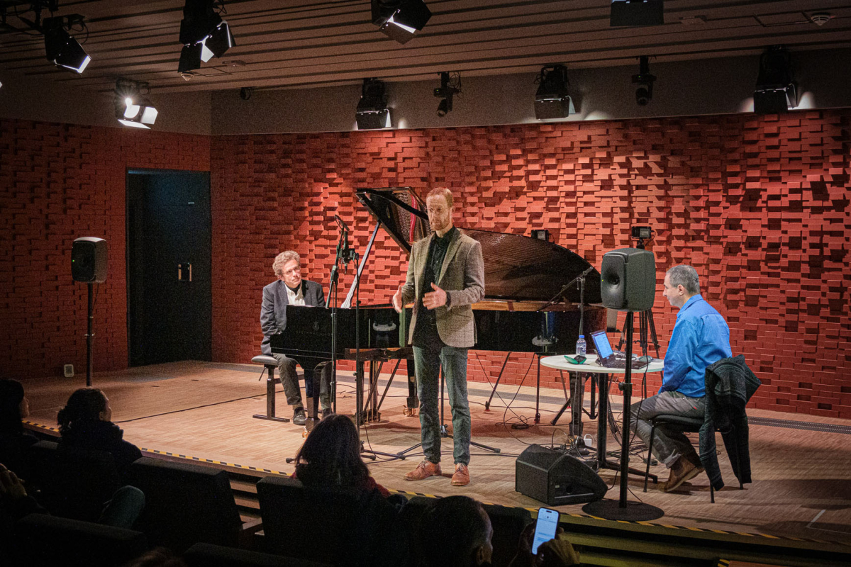 Eine Bühne, darauf ein Pianist an einem Flügel, ein Mensch an einem Mikrofon und ein Mensch an einem Tisch, darauf ein Laptop