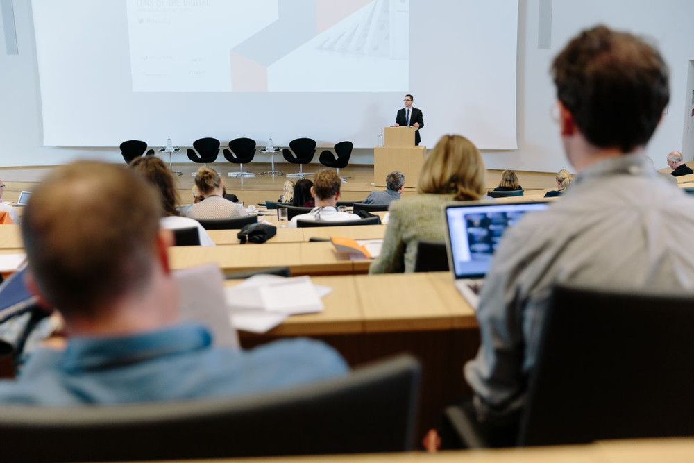 Menschen sind von hinten zu sehen. Sie sitzen in einem Auditorium wo eine Person am Podium steht.