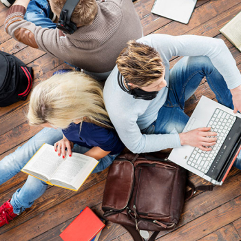 Das Foto zeigt zwei Personen von oben, wovon eine Person ein Buch liest und die andere am Laptop arbeitet.