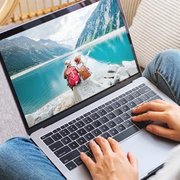 Person hat einen Laptop auf dem Schoß, auf dem man ein Bild mit zwei Personen am Bergsee sieht