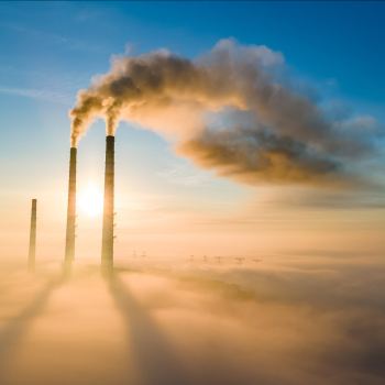 Das Bild zeigt drei industrielle Schornsteine, die Abgase vor einem Sonnenuntergang ausblasen.ist die EU-Flagge zu sehen,