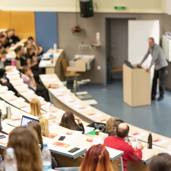 Das Bild zeigt einen Hörsaal mit Studierenden in den Reihen und einem Dozenten am Pult