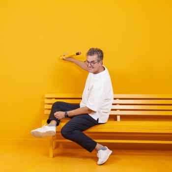 Das Bild zeigt Prof. Gunnar Spellmeyer kreativ auf einer Bank vor gelber Wand