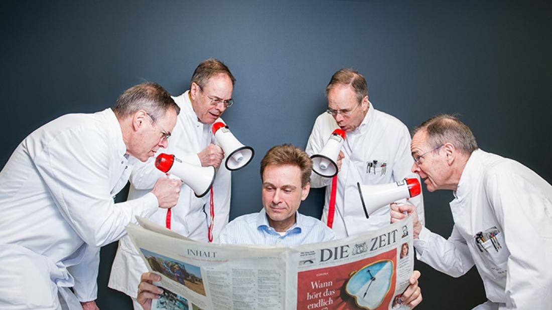 Ein Mann liest Zeitung und vier Personen in weißen Kitteln stehen mit Megaphonen um ihn herum