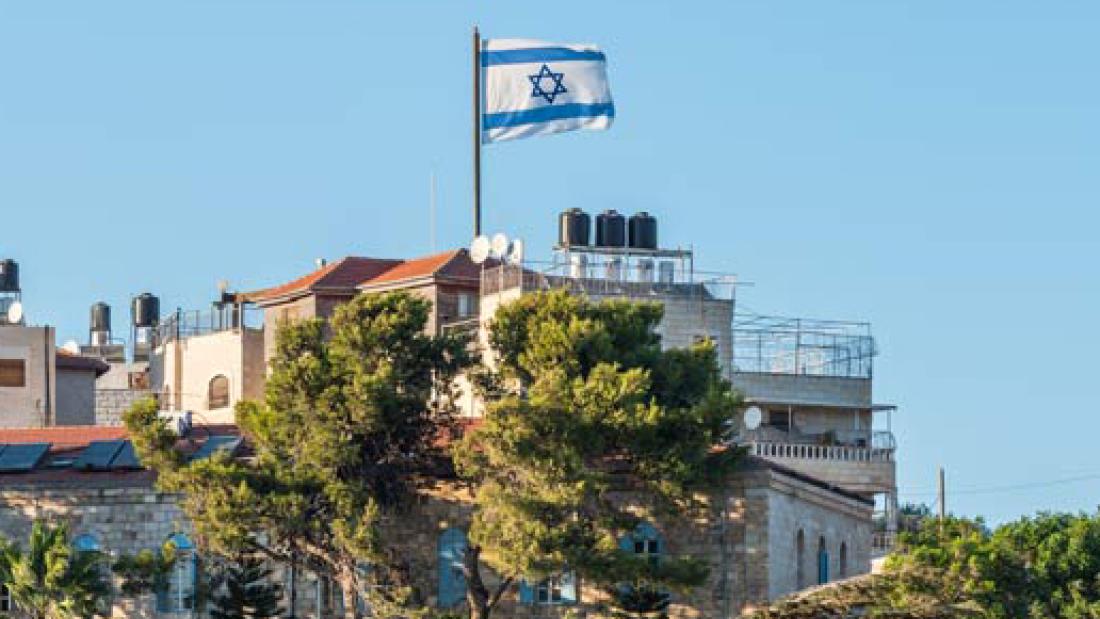 Eine Israel-Flagge weht über Wohengebäuden an einer felsigen Steilküste, die mit Bäumen bewachsen ist.