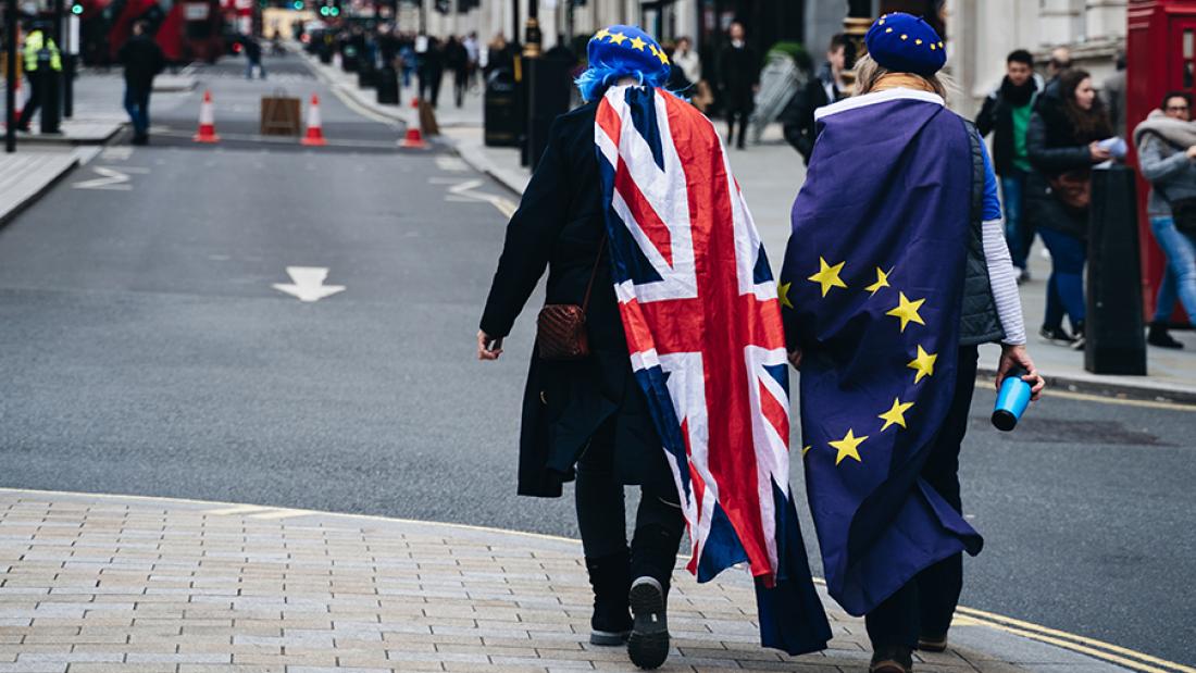 Zwei Menschen laufen durch eine Straße. Einer trägt den Union Jack, einer eine Europa-Flagge.