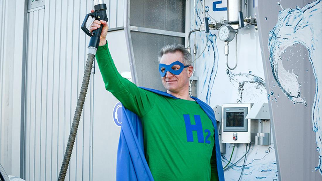 Richard Hanke-Rauschenbach trägt ein Heldenkostüm mit einem "H2"-Eblem und hält einen Tankrüssel für Wasserstoff in die Luft.