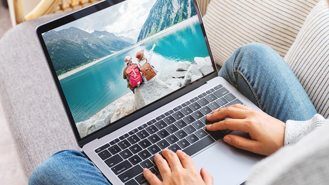 Mensch mit Laptop auf dem Schoß, der Bildschirm zeigt ein Foto mit zwei Rucksacktouristen vor schöner Landschaft