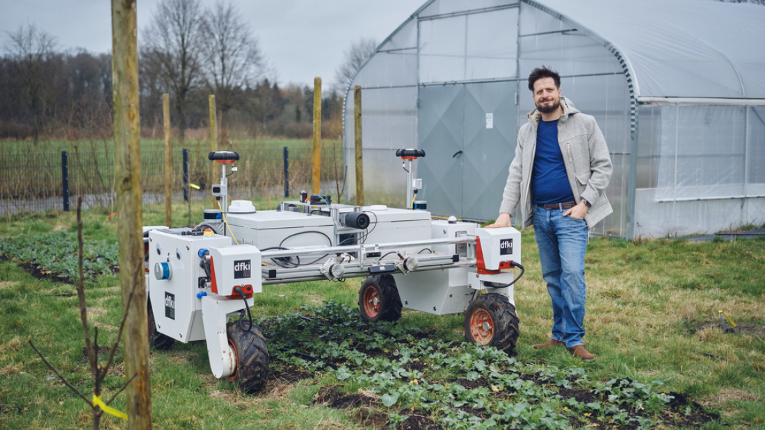 Ein Mann steht rechts neben einem weißen Roboter auf vier Rädern auf einem Feld